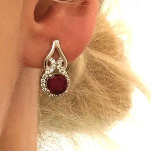May include: A silver-toned earring featuring a round, deep red gemstone at the center, surrounded by a halo of small, clear, sparkling stones. The setting has a teardrop-shaped frame that extends upward into a pointed, polished metal bail.