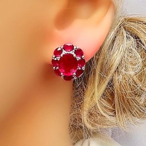 May include: A close-up of a stud earring with a large, oval, deep red gemstone at its centre, encircled by smaller, round red gemstones. The setting is silver or white gold, forming a floral pattern. The earring is on a person's ear.