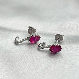 Puede incluir: Un par de pendientes de plata con un dise&ntilde;o curvo. Cada pendiente presenta una vibrante gema de rub&iacute; de forma ovalada y una fila de peque&ntilde;os acentos brillantes. Los pendientes se exhiben sobre una superficie blanca texturizada.