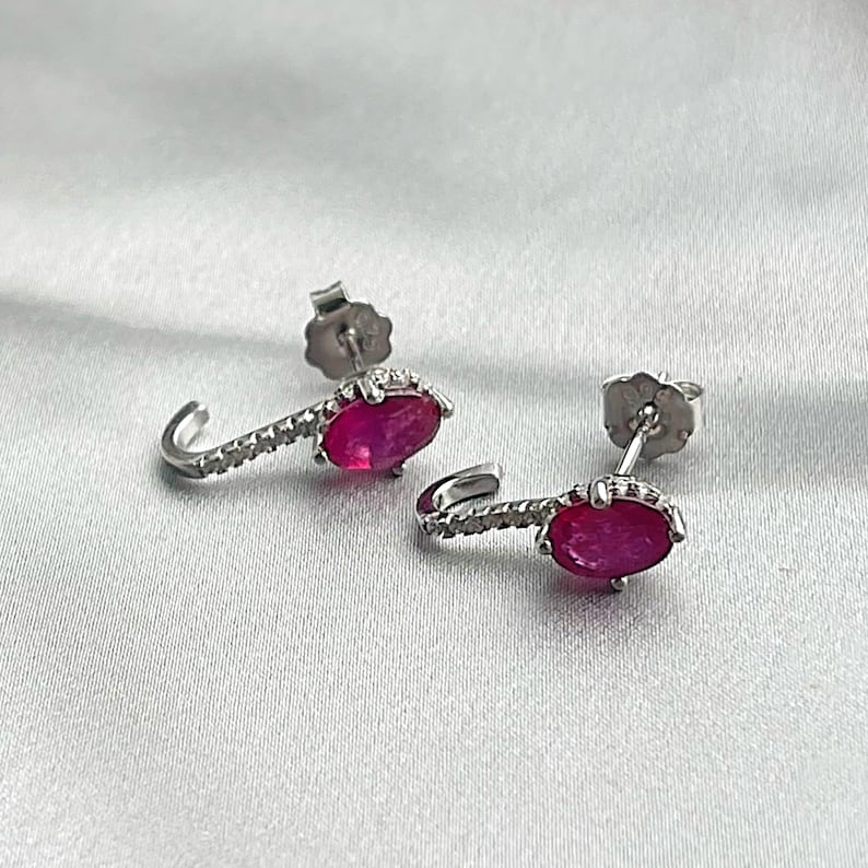 Puede incluir: Un par de pendientes de plata con un dise&ntilde;o curvo. Cada pendiente presenta una vibrante gema de rub&iacute; de forma ovalada y una fila de peque&ntilde;os acentos brillantes. Los pendientes se exhiben sobre una superficie blanca texturizada.