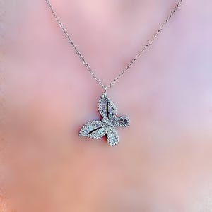 Puede incluir: Un collar con colgante de mariposa plateada. El dise&ntilde;o de la mariposa est&aacute; adornado con peque&ntilde;os cristales brillantes. La cadena del collar es delicada y plateada. El colgante est&aacute; centrado y el fondo es rosa p&aacute;lido.