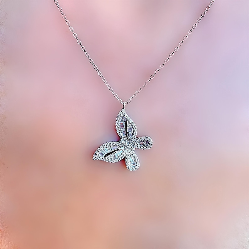 Puede incluir: Un collar con colgante de mariposa plateada. El dise&ntilde;o de la mariposa est&aacute; adornado con peque&ntilde;os cristales brillantes. La cadena del collar es delicada y plateada. El colgante est&aacute; centrado y el fondo es rosa p&aacute;lido.