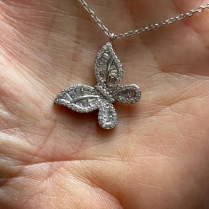 Puede incluir: Un collar con colgante de mariposa plateada. El dise&ntilde;o de la mariposa est&aacute; incrustado con piedras preciosas claras y brillantes. La cadena del collar es delicada y tambi&eacute;n plateada. El colgante se muestra sobre un fondo color piel.