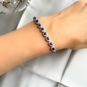 May include: A silver bracelet featuring a series of round, deep blue gemstones encircled by smaller, clear stones. The bracelet is displayed on a person's wrist against a soft, white background, showcasing its elegant design.