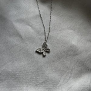 Puede incluir: Un collar con colgante de mariposa plateada. El colgante de mariposa est&aacute; incrustado con peque&ntilde;as piedras preciosas transparentes. La cadena del collar es delicada y plateada. El collar se muestra sobre un fondo de tela gris claro.