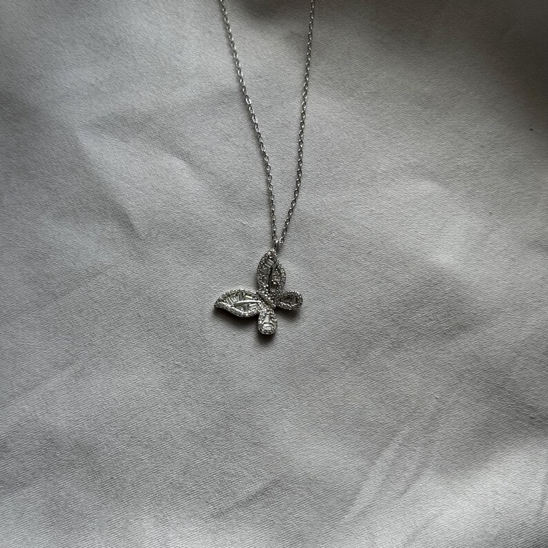 Puede incluir: Un collar con colgante de mariposa plateada. El colgante de mariposa est&aacute; incrustado con peque&ntilde;as piedras preciosas transparentes. La cadena del collar es delicada y plateada. El collar se muestra sobre un fondo de tela gris claro.