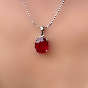 Könnte beinhalten: Eine silberne Halskette mit einem Anhänger, der einen großen, oval geschliffenen, tiefroten Edelstein zeigt. Der Edelstein ist in eine silberne Lünette gefasst und hängt an einer zarten Silberkette. Der Anhänger ist der Blickfang und zeigt die satte Farbe und den Glanz des Steins.