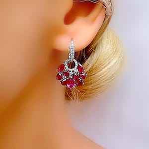 Könnte beinhalten: Nahaufnahme eines silbernen Ohrrings mit einem Cluster aus roten, tropfenförmigen Edelsteinen und kleinen weißen Steinen. Der Ohrring ist im Chandelier-Stil gestaltet.