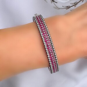 Puede incluir: Una pulsera plateada con una fila de pequeñas piedras rojas en el centro, flanqueada por filas de pequeñas cuentas plateadas redondas. La pulsera está en una muñeca, sobre un fondo de tela blanca.