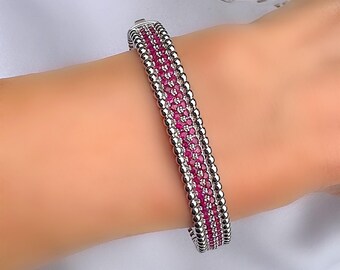 May include: A silver bracelet with a row of small, round silver beads on either side of a row of small, red gemstones. The bracelet is on a person's wrist, and the background is a soft white fabric.
