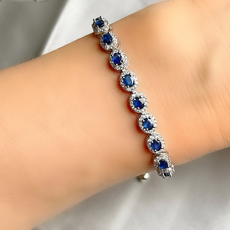 May include: A silver bracelet featuring oval-shaped, deep blue gemstones surrounded by smaller, clear stones. The bracelet is displayed on a person's wrist, showcasing its design and sparkle. The bracelet is a piece of jewelry.