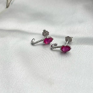 Puede incluir: Un par de pendientes plateados con un dise&ntilde;o curvo. Cada pendiente presenta una vibrante gema roja de forma ovalada y una fila de peque&ntilde;as piedras brillantes. Los pendientes se exhiben sobre una superficie blanca y sedosa.