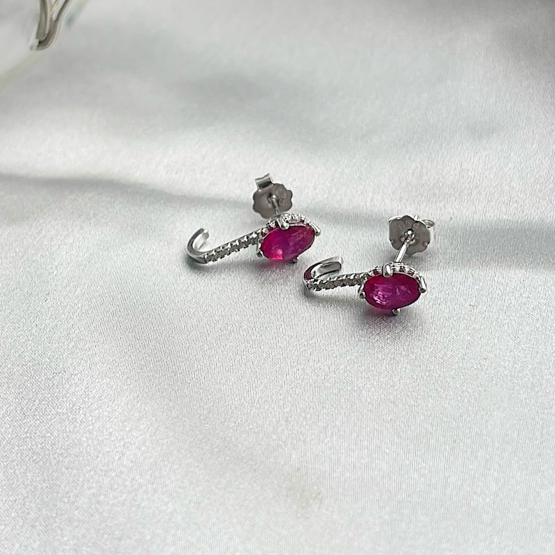 Puede incluir: Un par de pendientes plateados con un dise&ntilde;o curvo. Cada pendiente presenta una vibrante gema roja de forma ovalada y una fila de peque&ntilde;as piedras brillantes. Los pendientes se exhiben sobre una superficie blanca y sedosa.