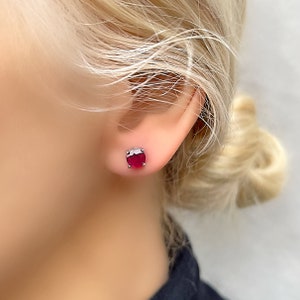 May include: A pair of silver stud earrings with a single oval-shaped ruby gemstone in each earring.