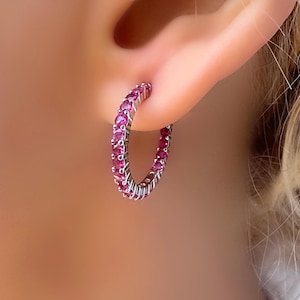May include: A silver hoop earring, set with a continuous line of small, round, pink gemstones. The earring is shown in an ear, with the skin tone visible in the background. The gemstones are closely set, creating a sparkling effect.