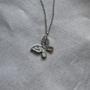 Puede incluir: Un collar con colgante de mariposa plateada. El colgante est&aacute; adornado con peque&ntilde;as piedras brillantes, formando una mariposa. La cadena del collar es fina y plateada, a juego con el dise&ntilde;o del colgante. El fondo es una tela gris claro.
