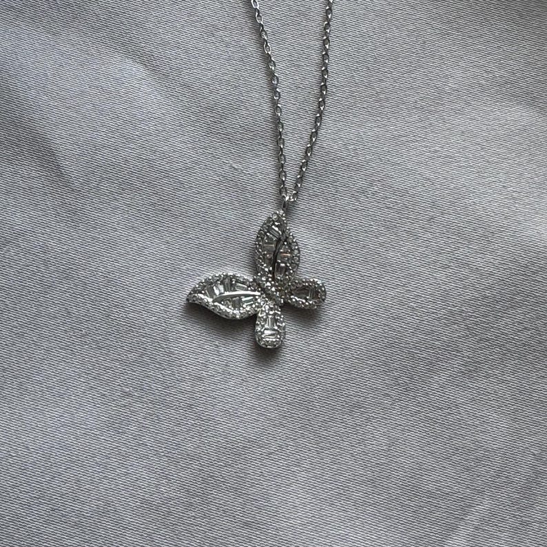 Puede incluir: Un collar con colgante de mariposa plateada. El colgante est&aacute; adornado con peque&ntilde;as piedras brillantes, formando una mariposa. La cadena del collar es fina y plateada, a juego con el dise&ntilde;o del colgante. El fondo es una tela gris claro.
