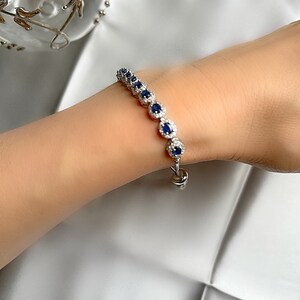 May include: A silver bracelet featuring a series of oval, dark blue gemstones surrounded by smaller, clear stones. The bracelet is displayed on a person's arm against a white background. The clasp is a simple hook and eye closure.