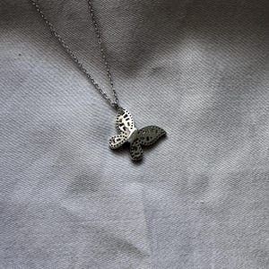Puede incluir: Collar con colgante de mariposa plateada. El colgante presenta un dise&ntilde;o detallado de mariposa con patrones recortados. El collar se muestra sobre un fondo de tela gris. La cadena es delicada y el colgante de mariposa es el punto focal.