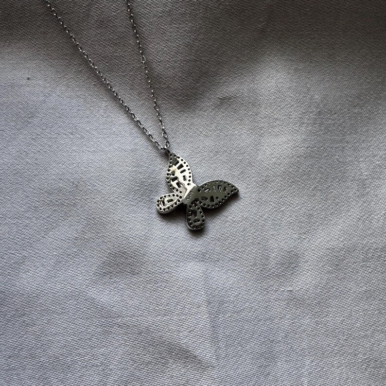 Puede incluir: Collar con colgante de mariposa plateada. El colgante presenta un dise&ntilde;o detallado de mariposa con patrones recortados. El collar se muestra sobre un fondo de tela gris. La cadena es delicada y el colgante de mariposa es el punto focal.