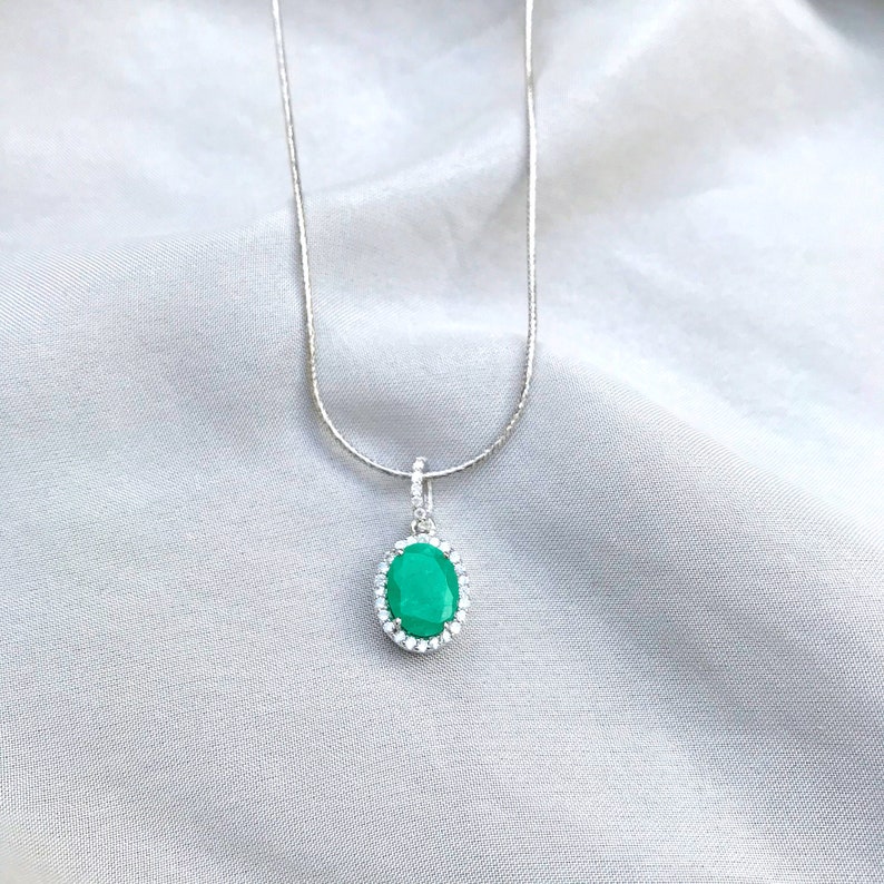 Puede incluir: Un collar de cadena de plata con un colgante que presenta una piedra preciosa verde de gran tama&ntilde;o de forma ovalada rodeada por un halo de piedras preciosas transparentes m&aacute;s peque&ntilde;as.