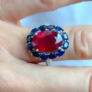 May include: A large, oval-shaped ruby ring with a halo of dark blue sapphires. The ring's band is silver and encrusted with small, clear gemstones. The ruby is a vibrant red color, and the sapphires are a deep blue.