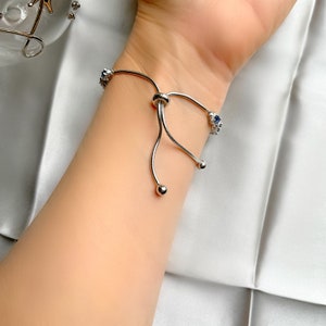 May include: A silver bracelet with blue gemstones is worn on a wrist. The bracelet has a delicate design with a sliding knot closure and small, round accents. The gemstones are set in a floral pattern.