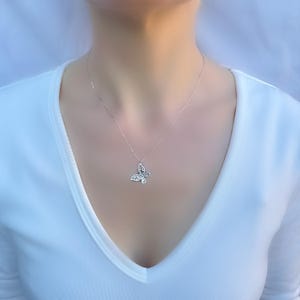 Puede incluir: Un collar con colgante de mariposa plateada. El colgante de mariposa est&aacute; adornado con peque&ntilde;as piedras brillantes. El collar se muestra sobre un fondo blanco, resaltando el dise&ntilde;o delicado y la cadena plateada.