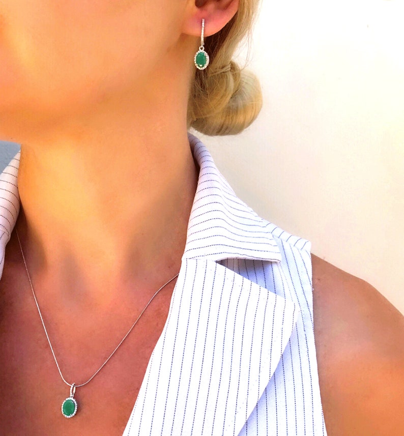 Puede incluir: Un collar y pendientes de plata con piedras preciosas verdes de forma ovalada. El collar tiene una cadena delicada y los pendientes son de estilo colgante.