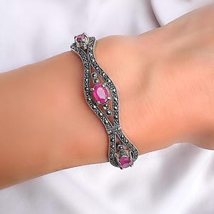 Puede incluir: Una pulsera articulada de tono plateado con un intrincado diseño calado acentuado con pequeñas piedras de color gris oscuro. La pieza está engastada con tres piedras preciosas ovaladas de color rojo rosado vibrante.