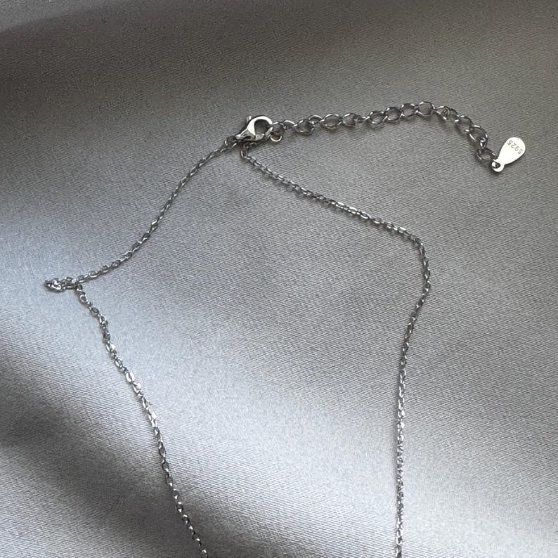 Puede incluir: Un collar plateado con una cadena delicada y una peque&ntilde;a etiqueta marcada "S925". El collar tiene un cierre de mosquet&oacute;n y una cadena ajustable. La joya se muestra sobre un fondo de sat&eacute;n plateado.