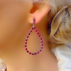 Op de afbeelding: Een paar druppelvormige oorbellen. Elke oorbel heeft een zilverkleurig metalen frame, dat een druppelvorm omlijnt. Het frame is versierd met talrijke kleine, levendige rode edelstenen, die een sprankelend effect creëren.