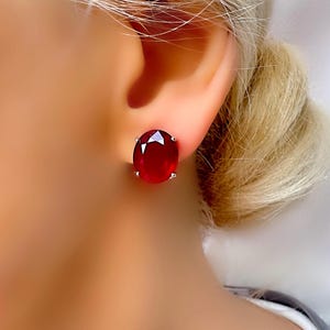 Peut inclure: Gros plan d'une boucle d'oreille à clou en forme de gemme rouge ovale. La pierre précieuse est d'un rouge profond et est sertie dans un métal de couleur argent. La boucle d'oreille est portée sur une oreille.