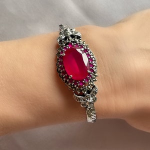 Könnte beinhalten: Ein silbernes Armreif-Armband mit einem großen ovalen roten Edelstein, umgeben von kleineren rosa Edelsteinen und silbernen Akzenten.