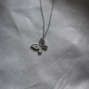 Puede incluir: Un collar con colgante de mariposa plateada. El colgante de mariposa est&aacute; incrustado con peque&ntilde;as piedras brillantes. La cadena del collar es fina y plateada. El fondo es una tela gris claro.
