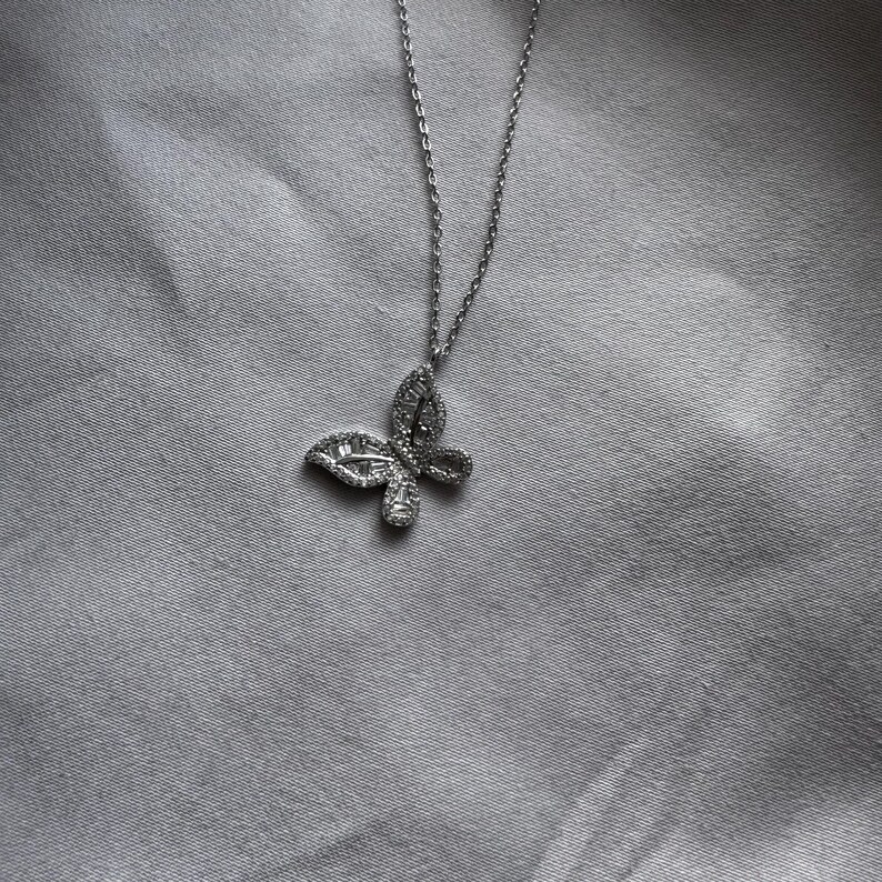Puede incluir: Un collar con colgante de mariposa plateada. El colgante de mariposa est&aacute; incrustado con peque&ntilde;as piedras brillantes. La cadena del collar es fina y plateada. El fondo es una tela gris claro.