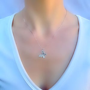 Puede incluir: Un collar con colgante de mariposa plateada. El colgante de mariposa est&aacute; adornado con peque&ntilde;as piedras brillantes. El collar lo lleva una persona que viste una camiseta blanca.