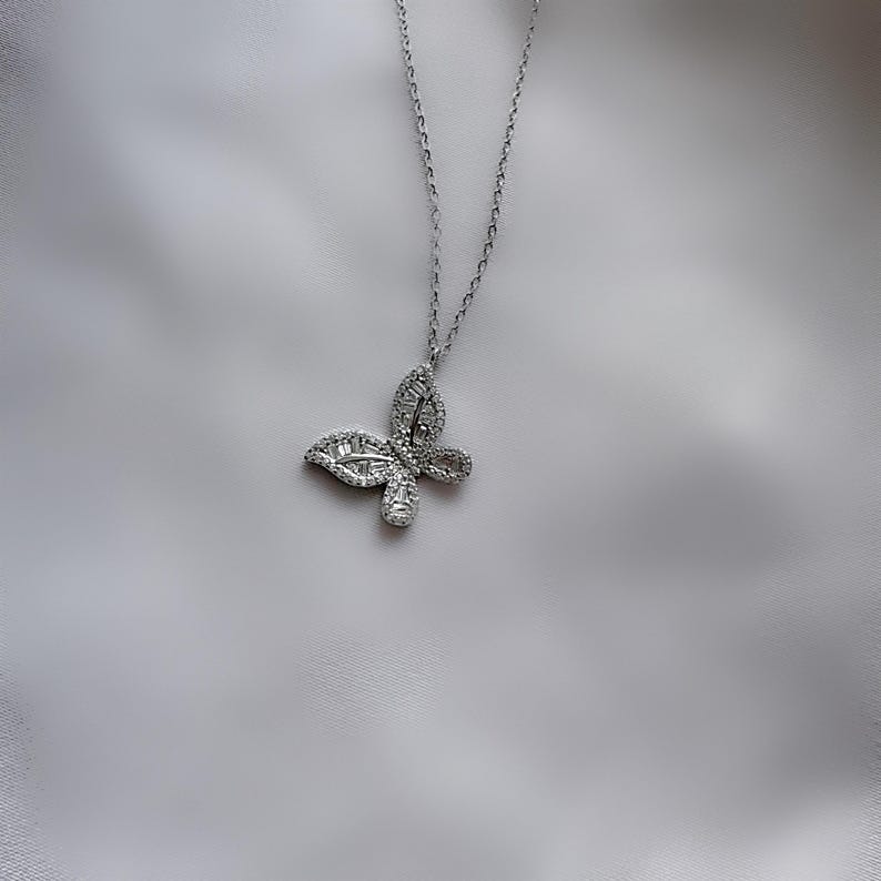 Puede incluir: Un collar con colgante de mariposa plateada. La mariposa est&aacute; adornada con peque&ntilde;as piedras brillantes. El collar cuelga de una delicada cadena plateada, creando una joya sencilla pero elegante.