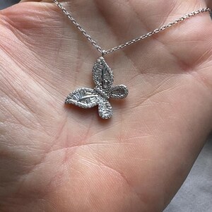 Puede incluir: Un collar con colgante de mariposa plateada. El dise&ntilde;o de la mariposa est&aacute; incrustado con peque&ntilde;as piedras brillantes. La cadena del collar es delicada y tambi&eacute;n plateada. El colgante est&aacute; centrado en la imagen.
