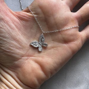 Puede incluir: Un collar con colgante de mariposa plateada. El colgante de mariposa est&aacute; adornado con peque&ntilde;as piedras brillantes. La cadena del collar es delicada y plateada. La joya se muestra sobre un fondo neutro.
