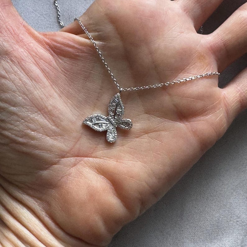 Puede incluir: Un collar con colgante de mariposa plateada. El colgante de mariposa est&aacute; adornado con peque&ntilde;as piedras brillantes. La cadena del collar es delicada y plateada. La joya se muestra sobre un fondo neutro.