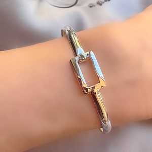 May include: A silver bangle bracelet with a gold square clasp. The clasp has a rectangular opening in the centre.