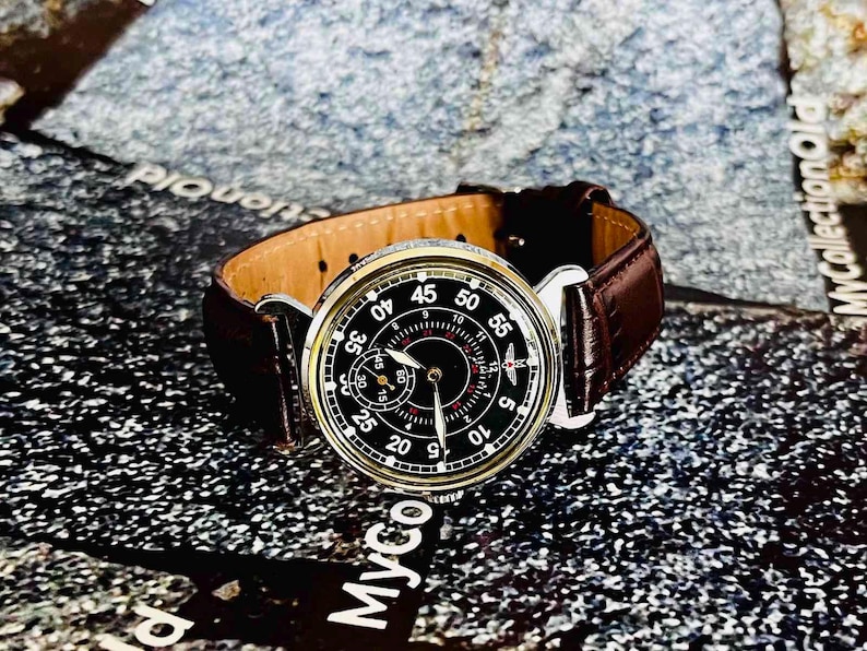May include: A vintage wristwatch with a black face, white numerals, and a brown leather band. The watch has a silver-colored case and a subdial. The band is dark brown and has a classic design.