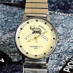 May include: Vintage wristwatch with a silver band and a cream-colored face. The watch face features a graphic of a lunar rover, Cyrillic text, and the text "LUNA-17" and "1970". The watch has gold-colored hands and black hour markers.