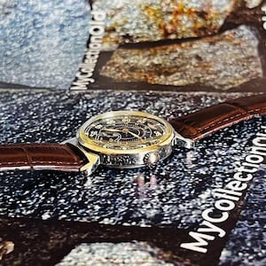May include: A vintage wristwatch with a black face, gold-colored bezel, and brown leather band. The watch features a detailed dial with numerals and hands. The band has a textured, crocodile-like pattern. The watch is displayed on a textured surface.