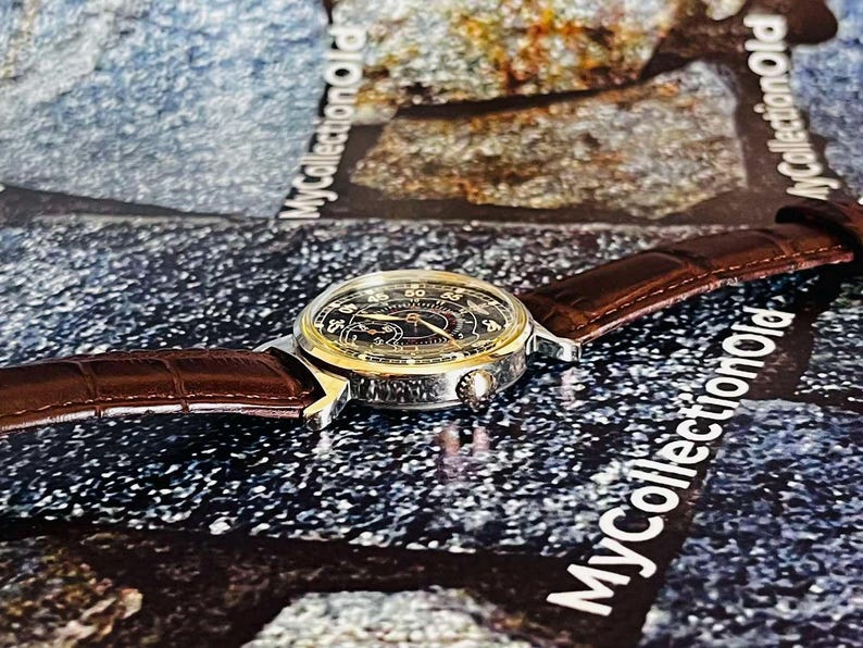 May include: A vintage wristwatch with a black face, gold-colored bezel, and brown leather band. The watch features a detailed dial with numerals and hands. The band has a textured, crocodile-like pattern. The watch is displayed on a textured surface.