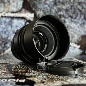 Puede incluir: Un objetivo de cámara negro con una tapa de lente. El objetivo tiene un anillo plateado con el texto "FE 50mm F1.4 GM" impreso en él.