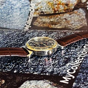 May include: A vintage wristwatch with a black face, gold-colored bezel, and brown leather band. The watch features a detailed dial with numerals and hands. The band has a textured, crocodile-like pattern. The watch is displayed on a textured surface.