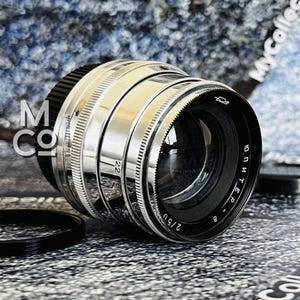 Può includere: Un obiettivo per fotocamera vintage con finitura argento e nera. L'obiettivo presenta una serie di anelli e marcature, tra cui il numero "22" e il testo "2/50". Sono visibili anche un tappo dell'obiettivo nero e un anello dell'obiettivo.