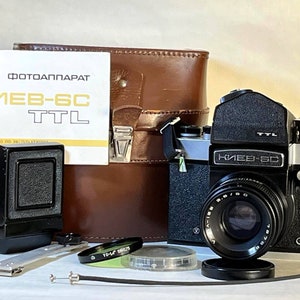 May include: A black and silver Kiev-6C TTL camera with a lens attached. The camera is sitting on a white surface with a brown leather case, a black camera back, and a lens filter.